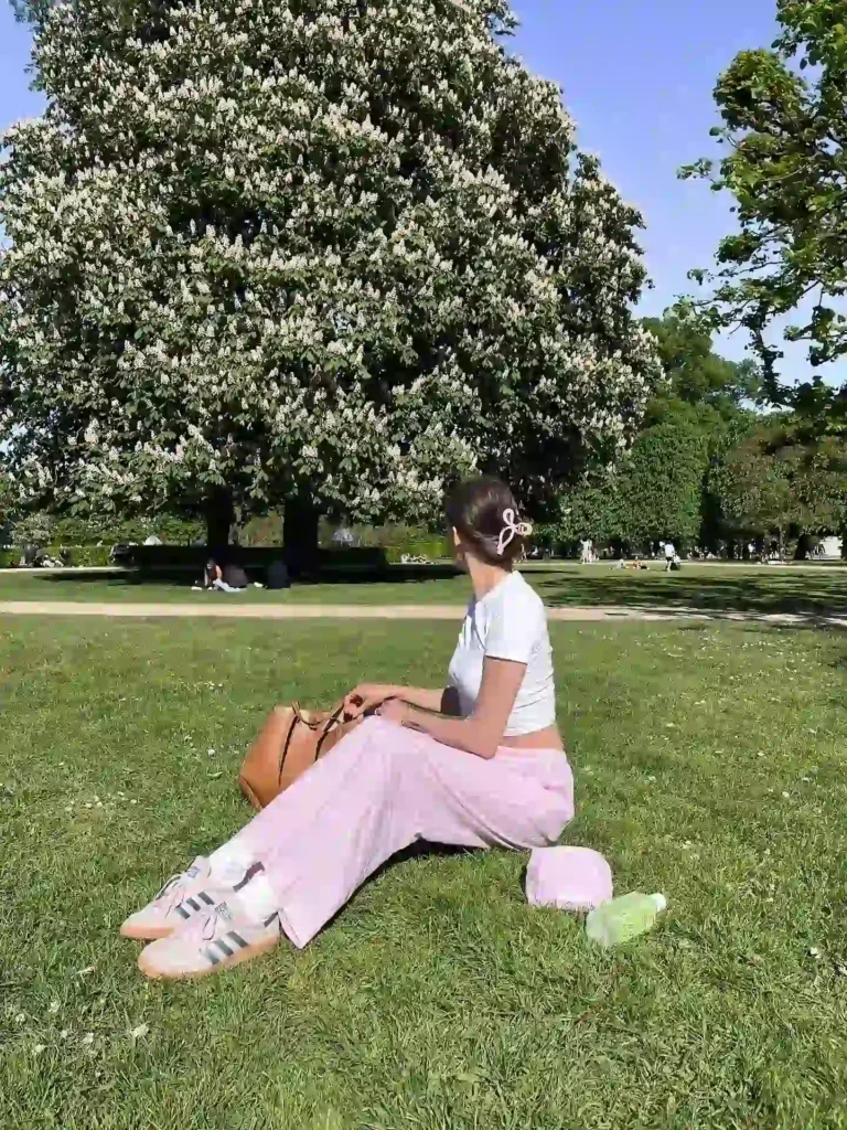 Woman seated on grass in a park, wearing a white top and pink pants, enjoying a sunny day under a flowering tree.