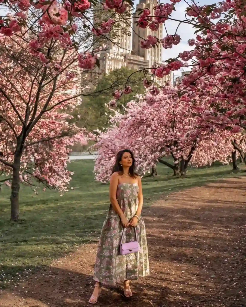 Woman in floral dress stands amidst blooming cherry blossom trees in a park with a cityscape in the background.