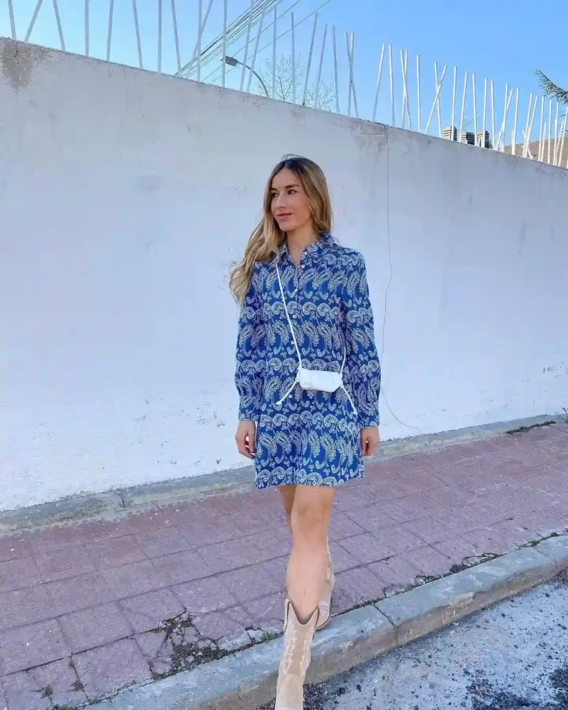 Woman in a blue paisley dress and beige cowboy boots walking along a sidewalk next to a white wall under a clear sky.