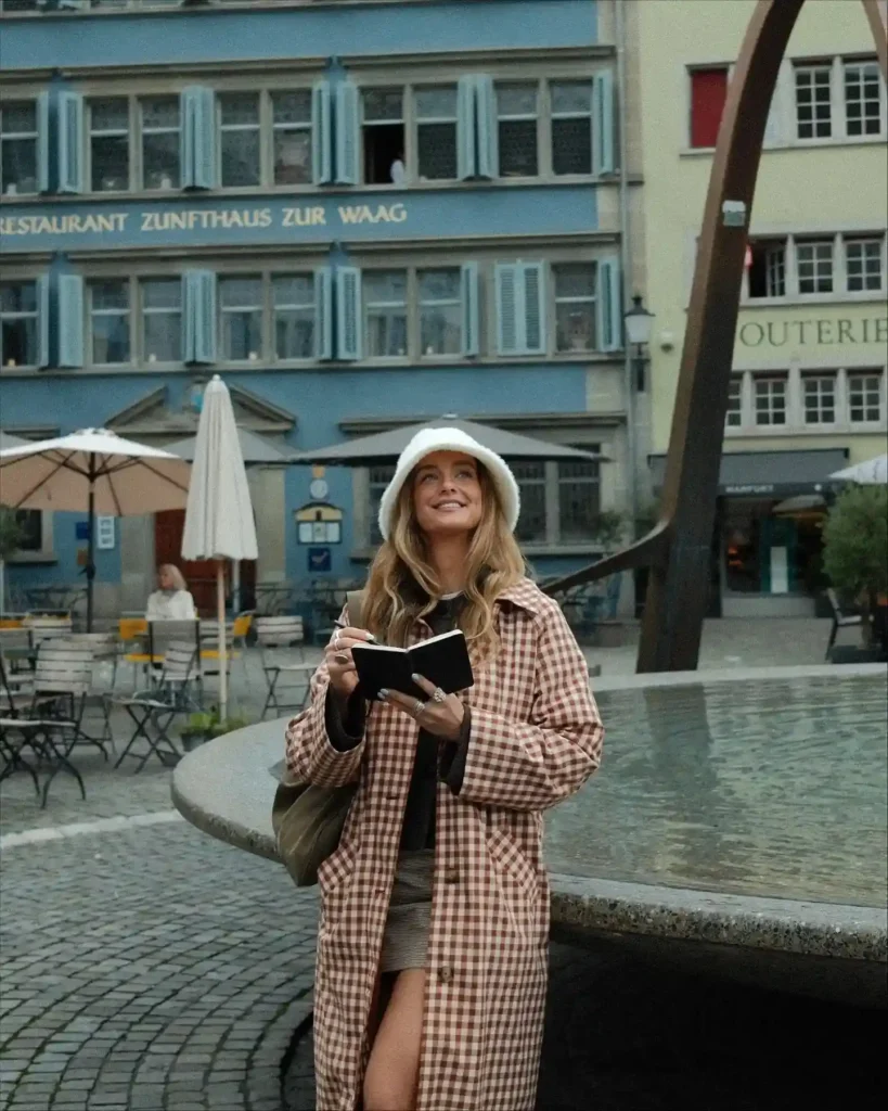 Woman in a checkered coat smiles while writing in a notebook by a fountain in a charming city square.