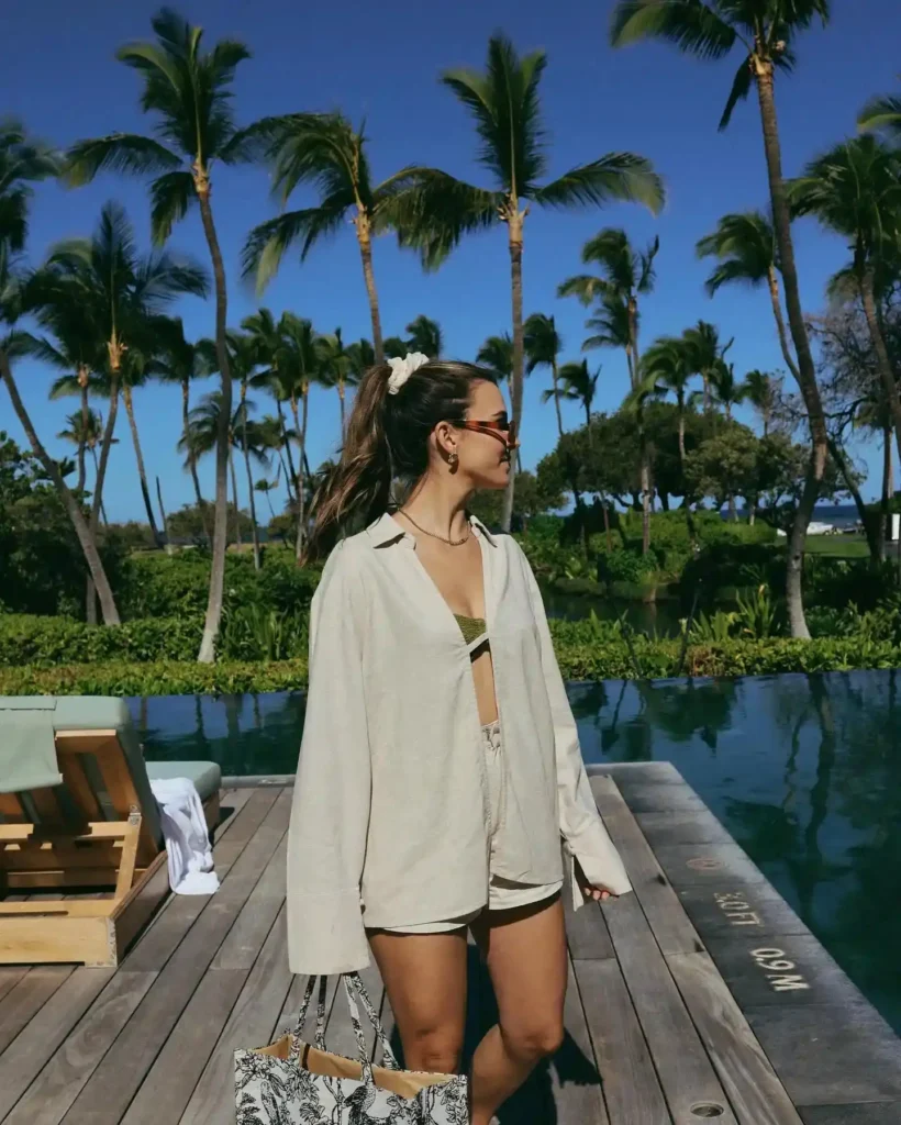 A woman in a stylish outfit stands by a scenic pool surrounded by palm trees, enjoying a sunny day outdoors.