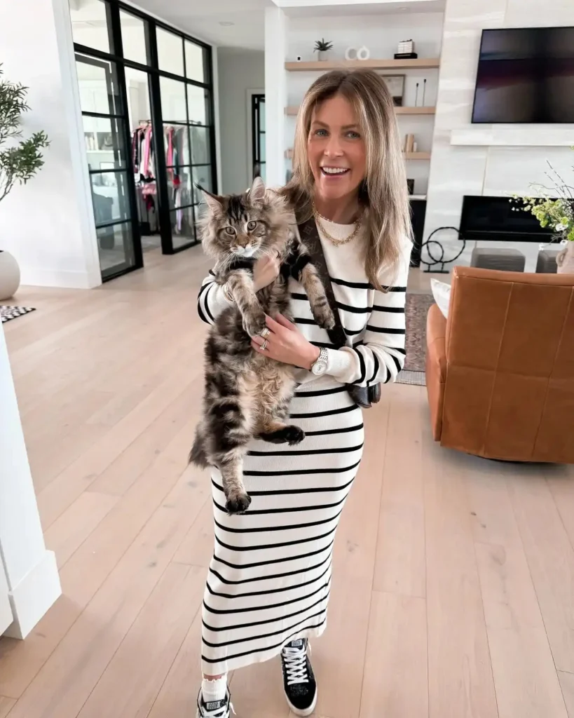 Smiling woman in a striped dress holding a fluffy cat in a stylish, modern living room.