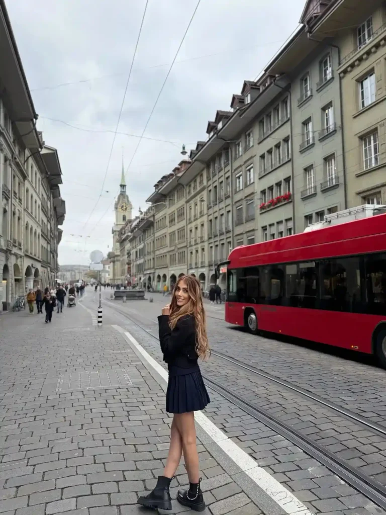 Young woman in a black outfit smiles while standing on a cobblestone street in Bern, Switzerland, with a red bus nearby.