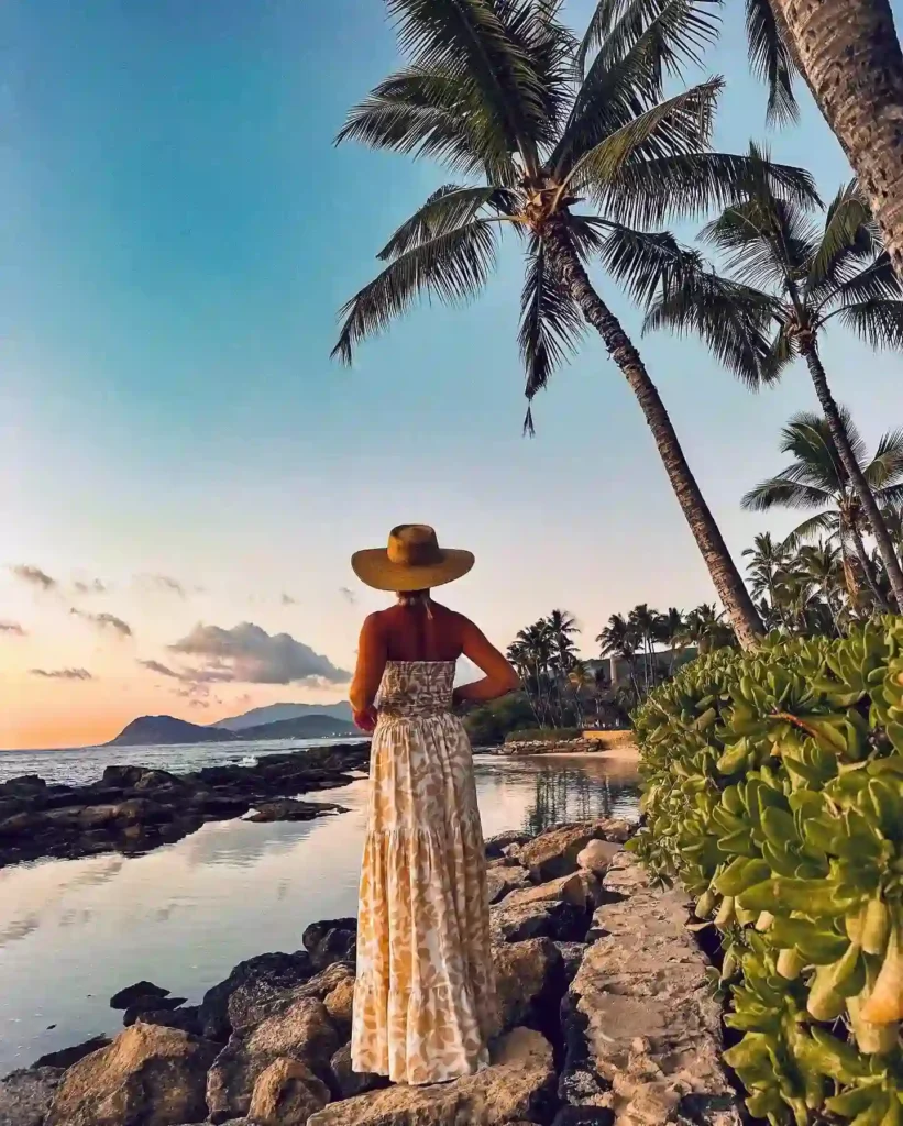 Woman in a flowing dress and hat stands by a serene beach at sunset, surrounded by palm trees and rocks.