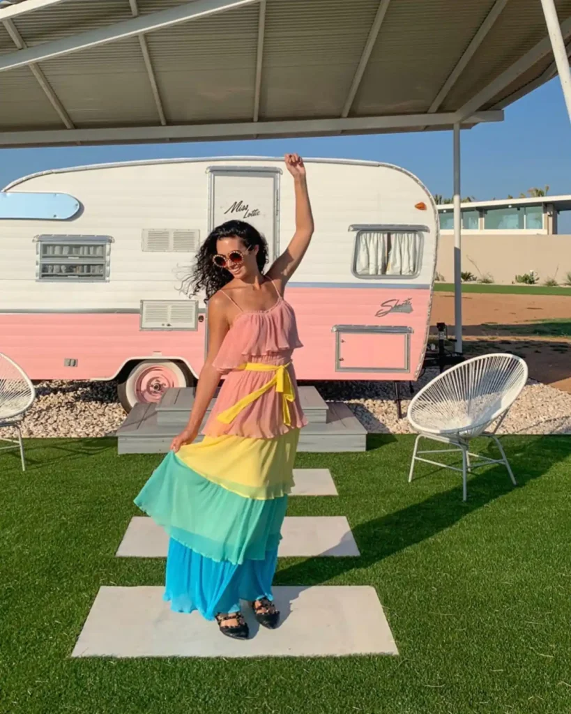 Woman posing playfully in a colorful layered dress near a vintage pink camper in a sunny outdoor setting.