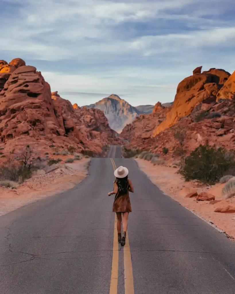 A woman walks along a scenic road surrounded by stunning red rock formations and mountains under a blue sky.