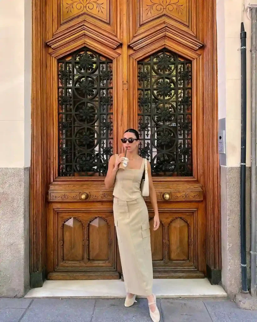 Woman in stylish beige dress sipping coffee, standing in front of an ornate wooden door with decorative ironwork.