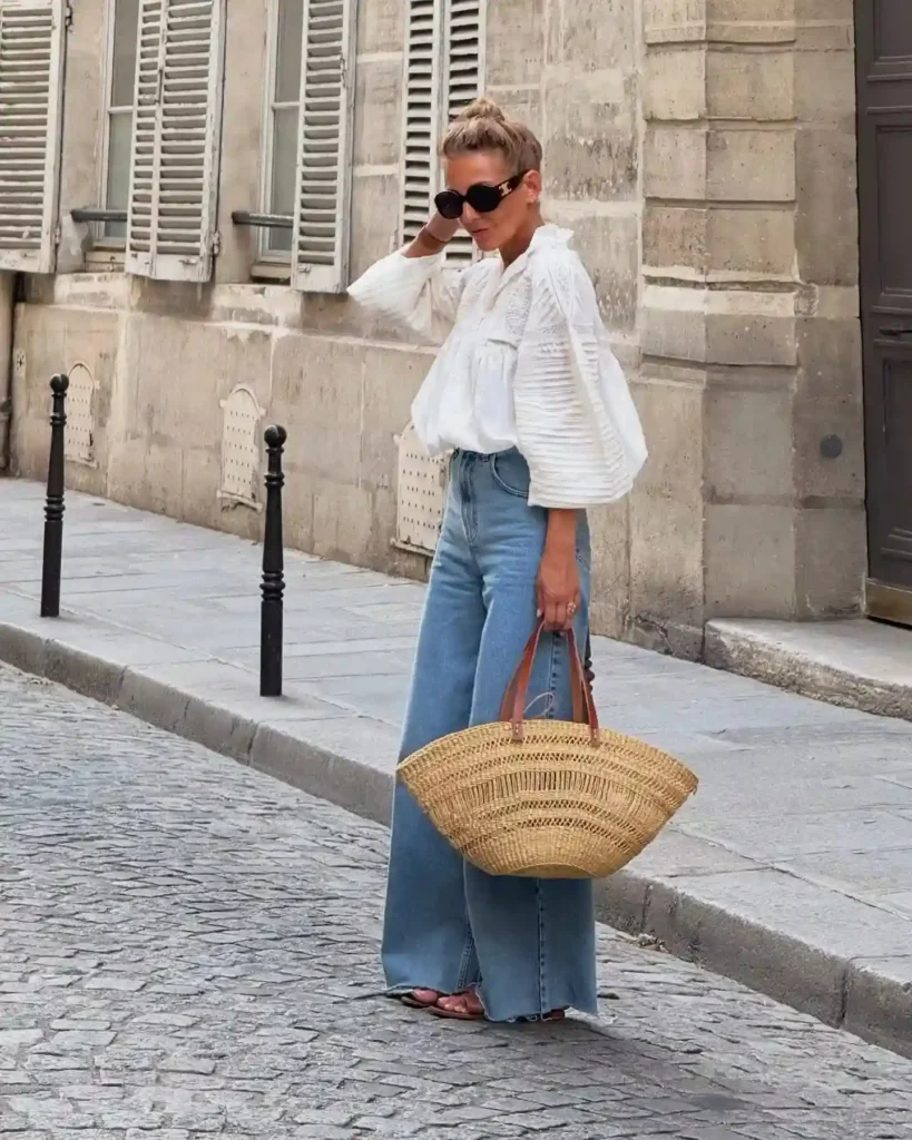 Stylish woman in a white blouse and wide-leg jeans, holding a straw bag, poses on a cobblestone street in Paris.