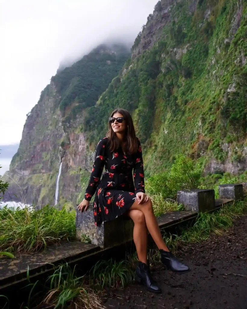 Young woman in sunglasses seated on a stone ledge, surrounded by lush green mountains and a waterfall in the background.