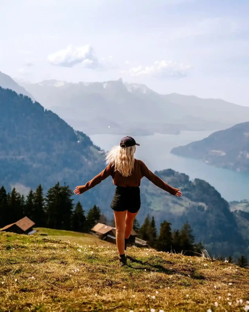 Woman in a brown sweater and cap joyfully exploring a scenic mountain view, surrounded by lush greenery and a lake.