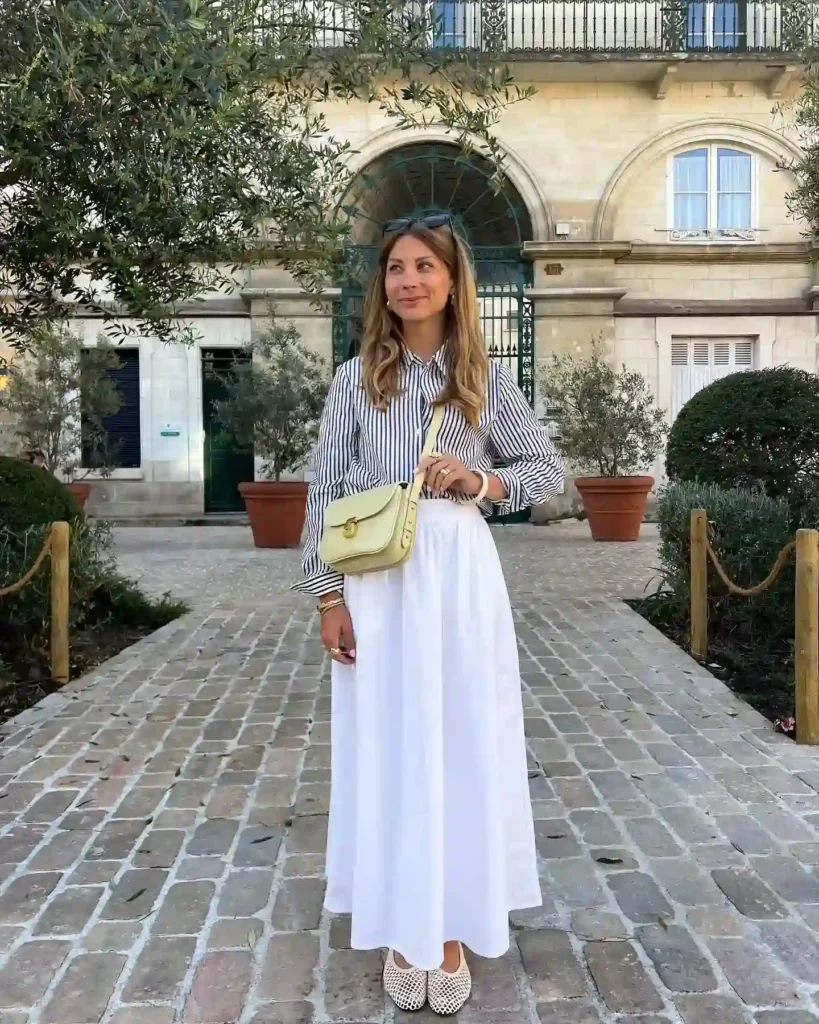 Stylish woman in a striped shirt and white skirt, posing outdoors with greenery and a historic building in the background.