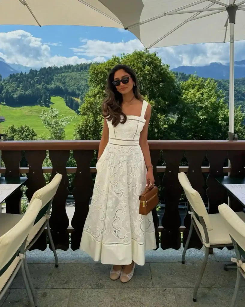 A woman stands on a balcony in a white dress, sunglasses on, with a scenic mountain view in the background.