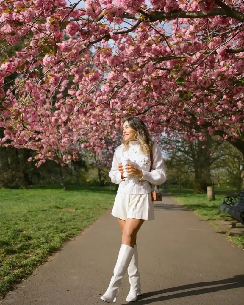 Woman in a white sweater and skirt stands under blooming cherry blossom trees, holding a drink in springtime park.