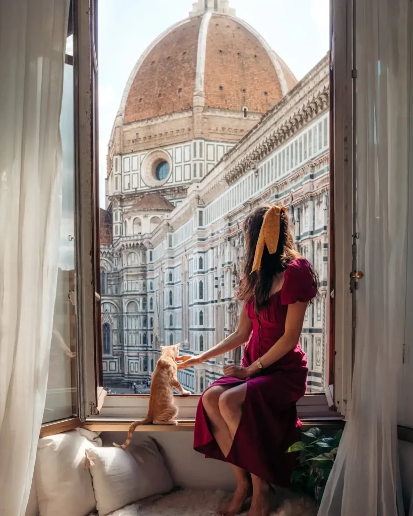 Woman in a red dress enjoying the view of Florence, Italy, with her orange cat at an open window.