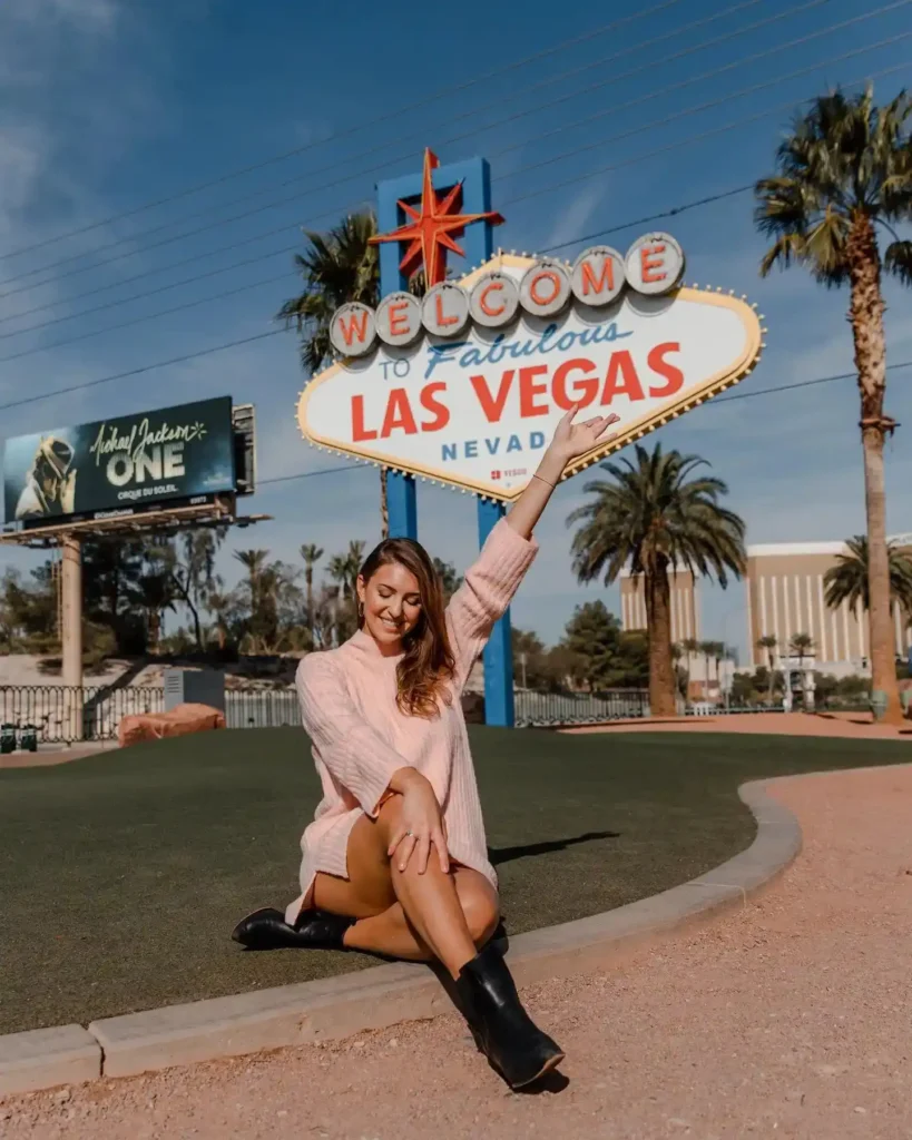 Woman in a pink sweater poses at the iconic Las Vegas sign, surrounded by palm trees under a clear blue sky.