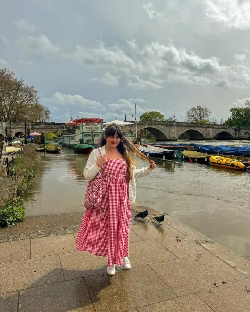 Young woman in a red gingham dress by a river with boats, enjoying a cloudy day outdoors.