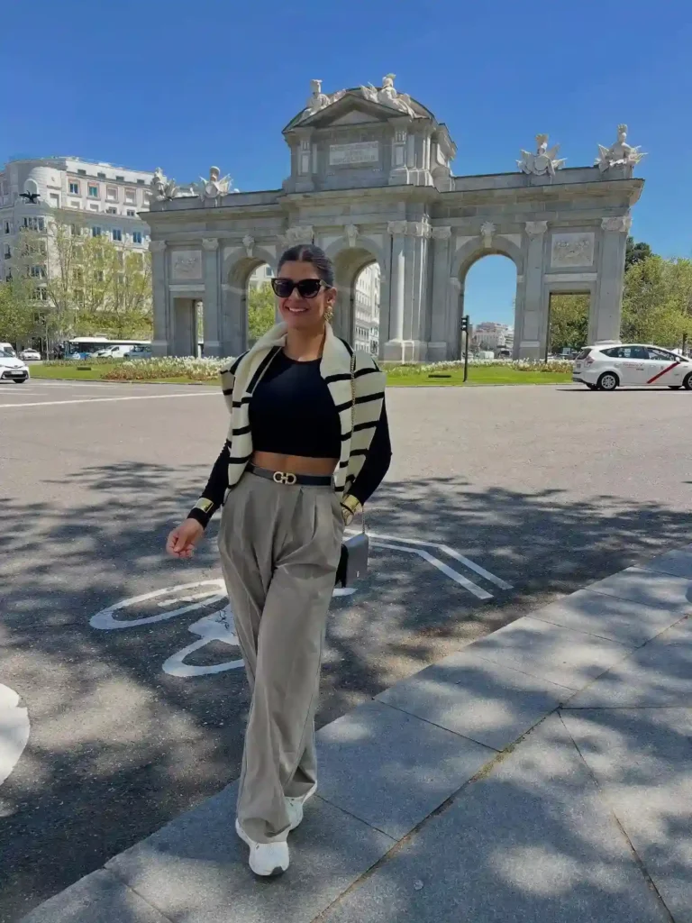 Stylish woman in fashionable attire posing in front of a historic archway on a sunny day.