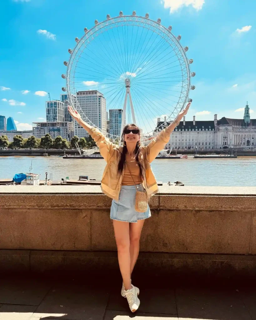 A woman smiles with arms raised by the Thames River, the London Eye towering in the background on a sunny day.