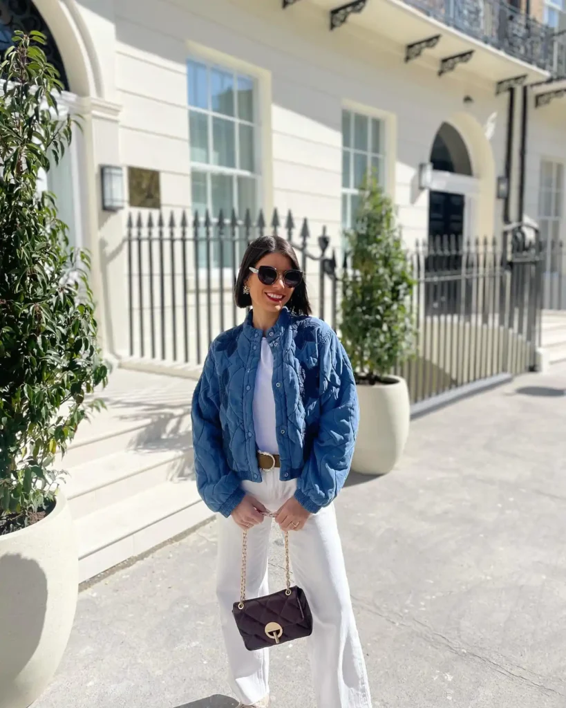Stylish woman in a blue quilted jacket and white pants, posing outside a chic building with sunlight and greenery.