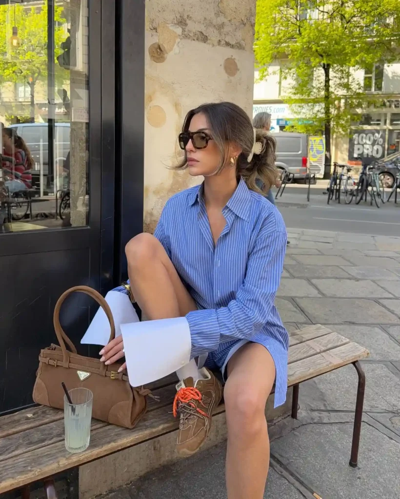 Woman in oversized striped shirt and sunglasses sits on a bench, holding a handbag, with a drink beside her.