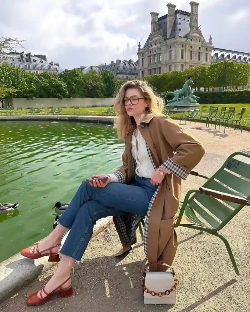 A woman in a stylish outfit sits by a green pond, enjoying a scenic view of trees and classic architecture.