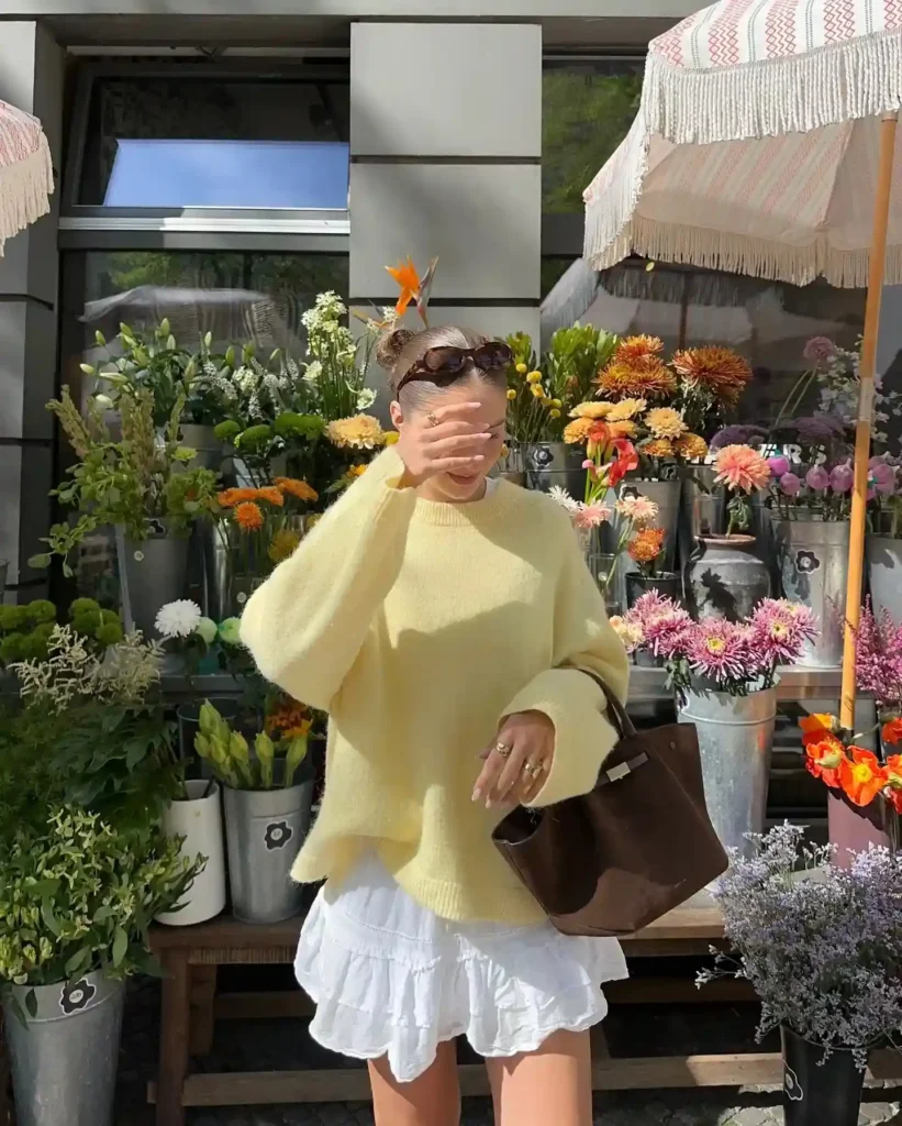 Woman in a yellow sweater and white skirt poses in front of a colorful flower shop, holding a bag and shielding her eyes.