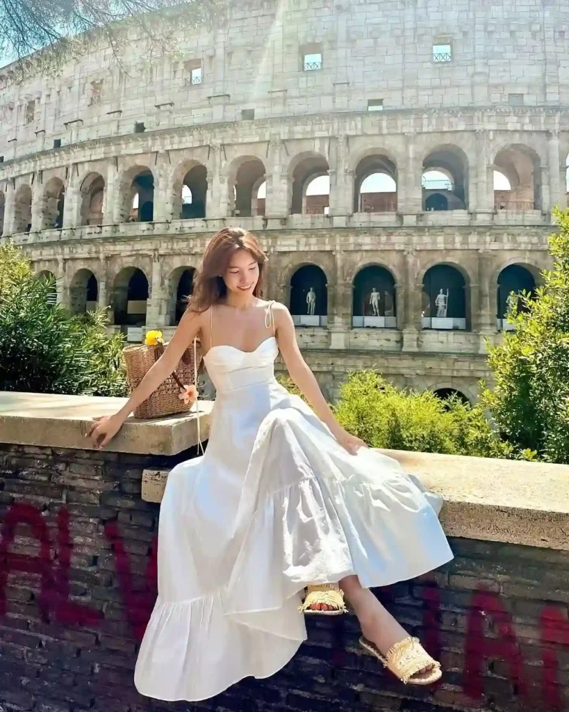 Young woman in a white dress sitting on a wall with the Colosseum in the background, enjoying a sunny day in Rome.