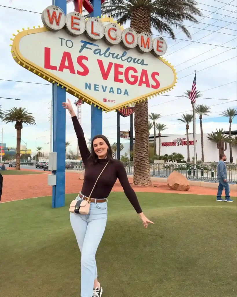 A cheerful traveler poses at the iconic Welcome to Las Vegas sign, showcasing palm trees and vibrant surroundings.