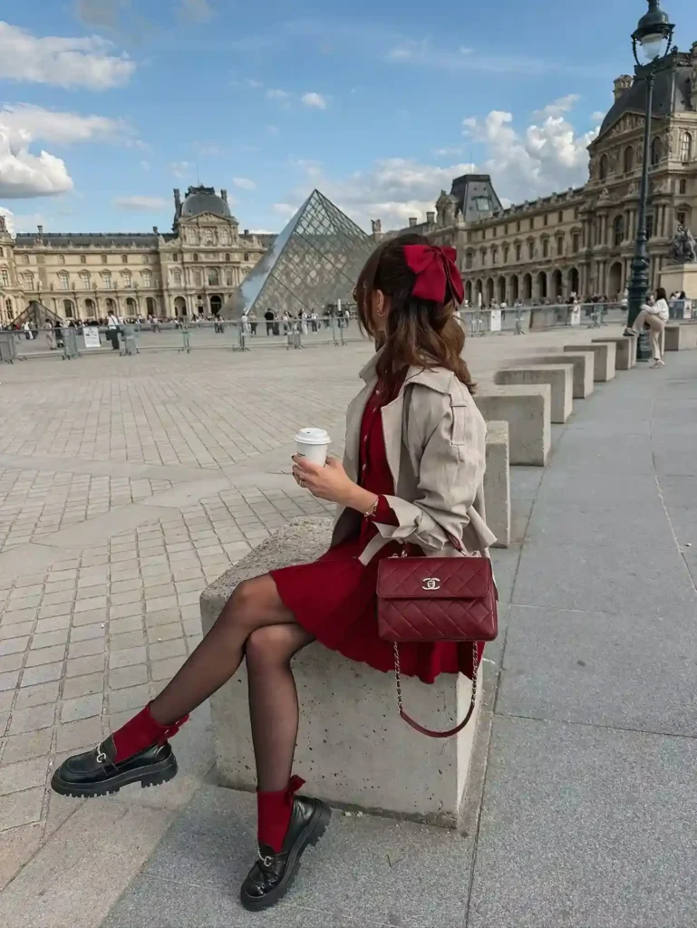 Fashionable woman in red dress and stylish black shoes enjoying coffee at the Louvre, Paris, with iconic pyramid in background.
