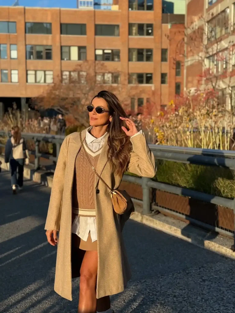 Woman in a stylish tan coat and sunglasses poses outdoors in a sunlit urban setting, showcasing a chic fall outfit.