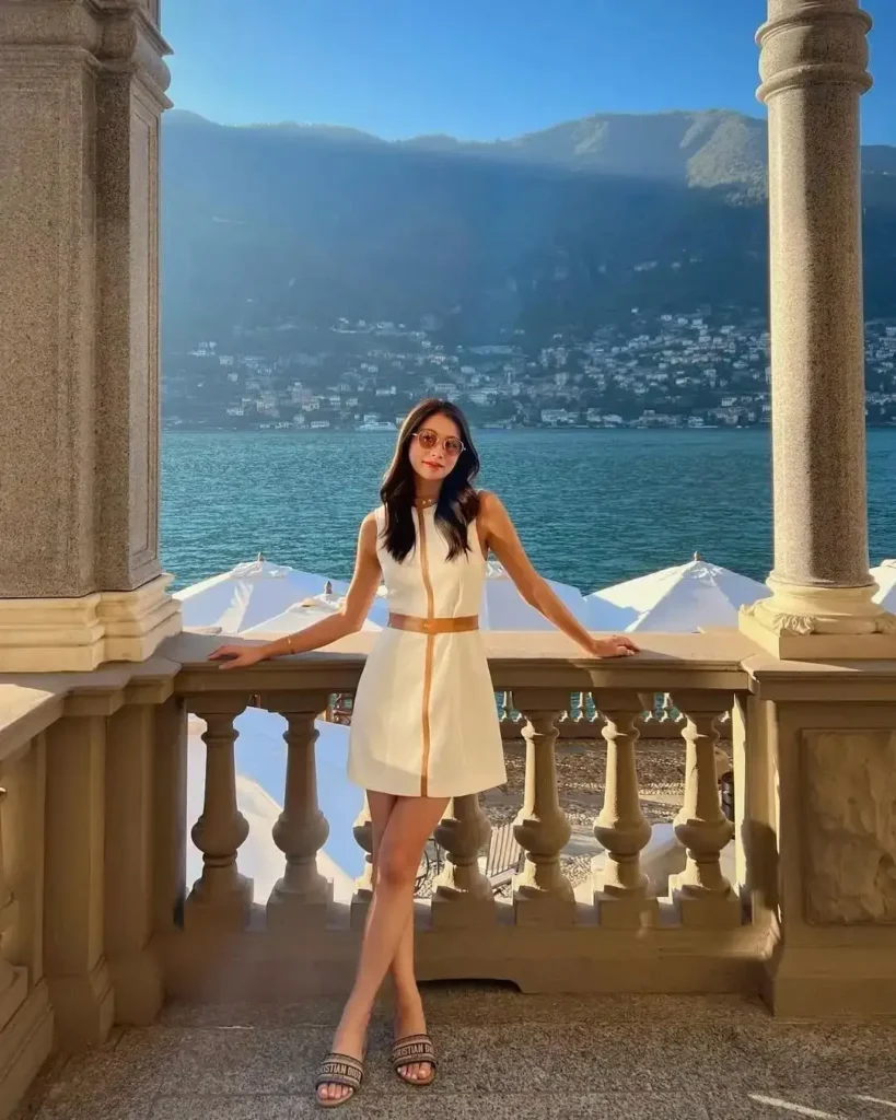 A woman in a white dress poses by a scenic lake, showcasing elegant architecture and lush mountains in the background.