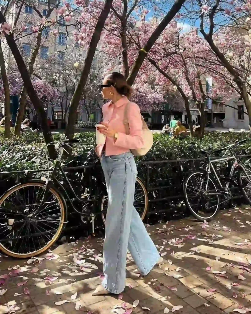 Young woman in pink jacket and wide jeans strolls among blooming cherry trees and fallen petals in urban park.