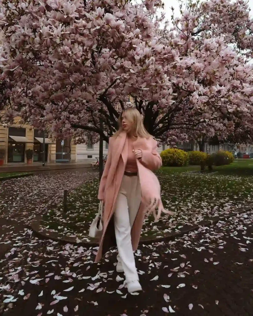 Woman in a stylish pink coat walks among blooming magnolia trees, surrounded by fallen petals on a scenic urban path.