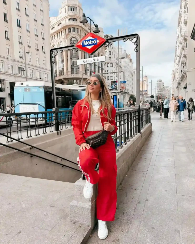 Young woman in red outfit poses by Gran Via Metro sign in a bustling city street, showcasing urban travel style.