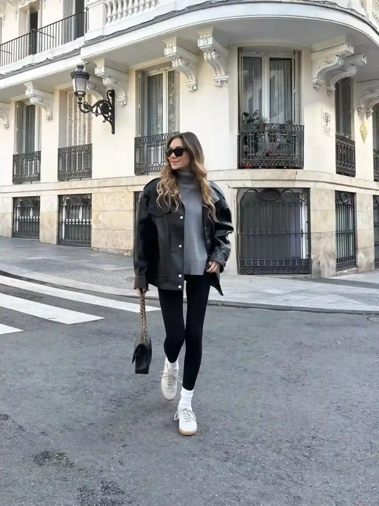 Stylish woman in a black leather jacket and leggings walks confidently along a city street with elegant architecture.