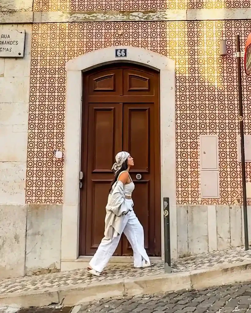 A woman in stylish white attire walks past a colorful tiled wall and a wooden door in a charming urban setting.