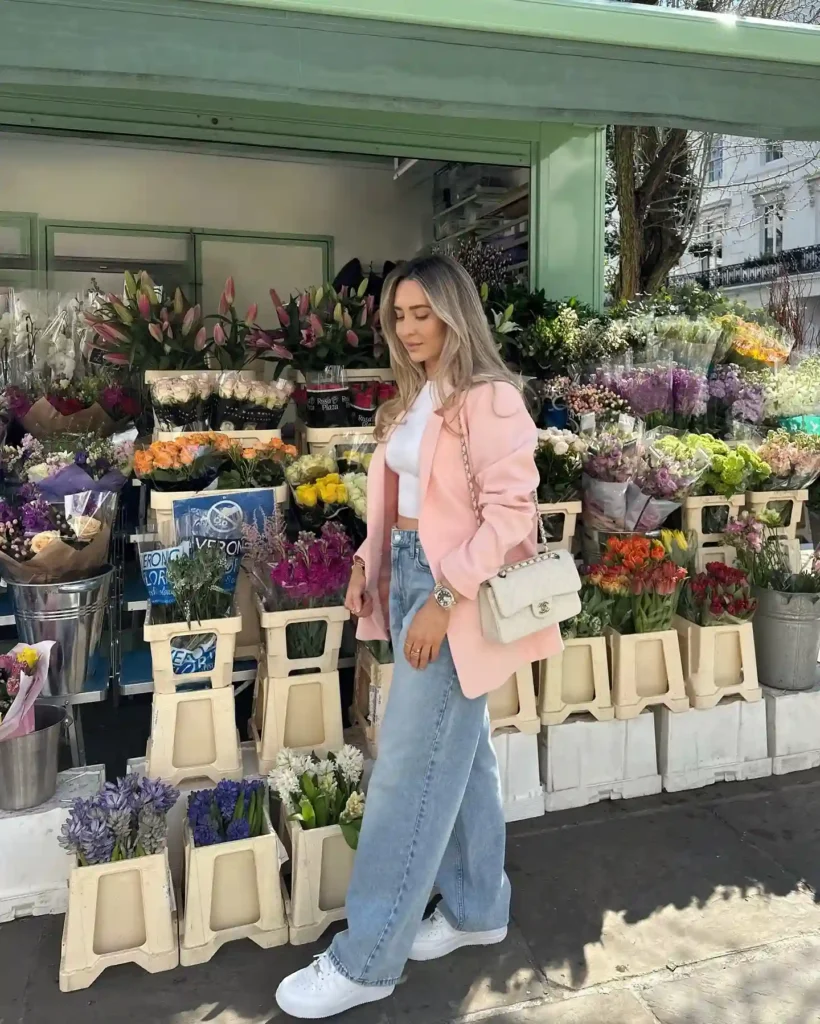 Young woman in a pastel pink blazer stands in front of a vibrant flower shop, surrounded by colorful blooms.