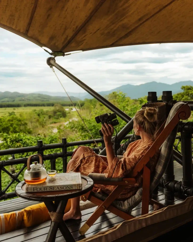 Woman relaxing in a hammock, holding a camera, with a scenic view of mountains and greenery. A teapot and book are nearby.