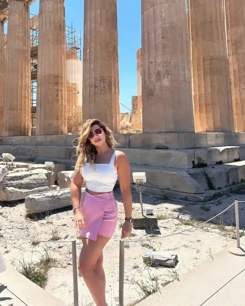 Young woman in a white top and pink skirt stands near ancient columns under a clear blue sky. Historical site backdrop.