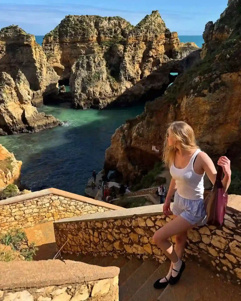 A woman in casual attire walks down stone steps overlooking a scenic coastal landscape with rocky cliffs and turquoise water.