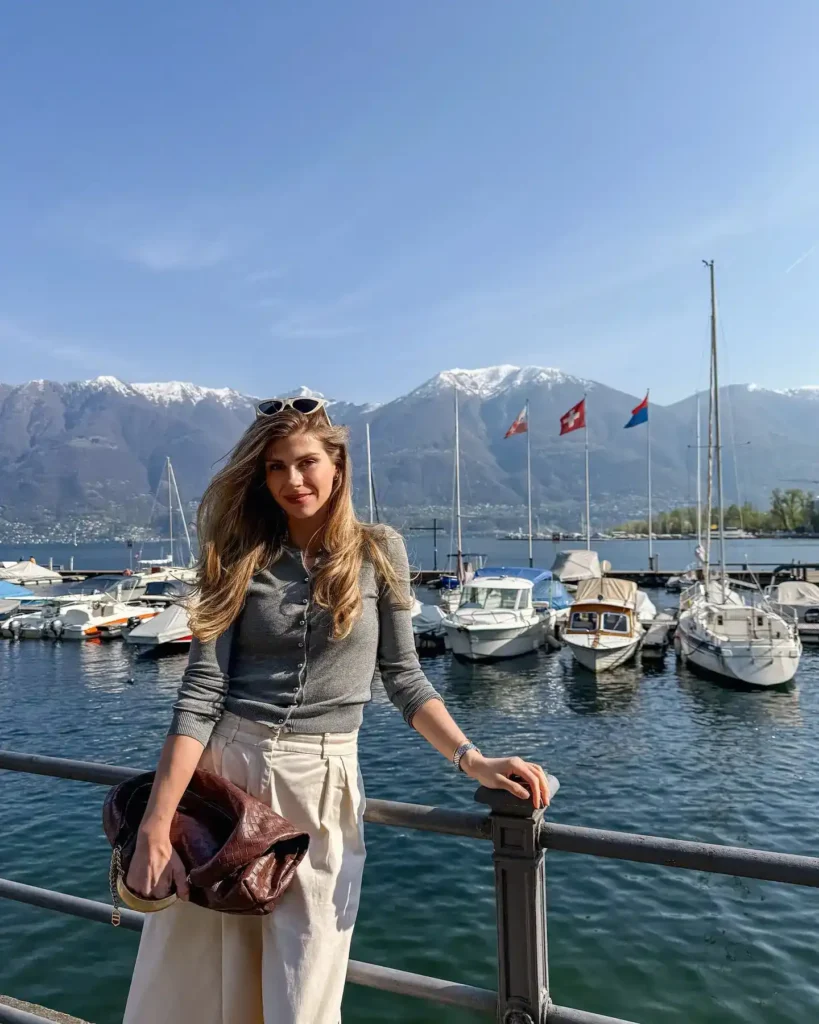 Woman standing by a marina with boats, mountains, and blue sky in the background, showcasing a sunny day.