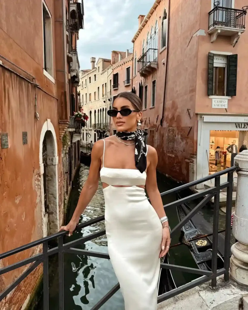 Stylish woman in a white dress and sunglasses poses on a canal bridge in Venice, surrounded by historic buildings.