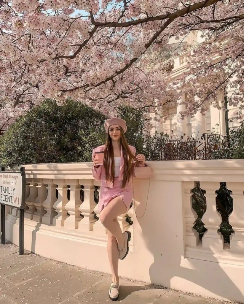 Fashionable woman in pink outfit poses near blossoming cherry trees in a sunny park, sipping a drink.