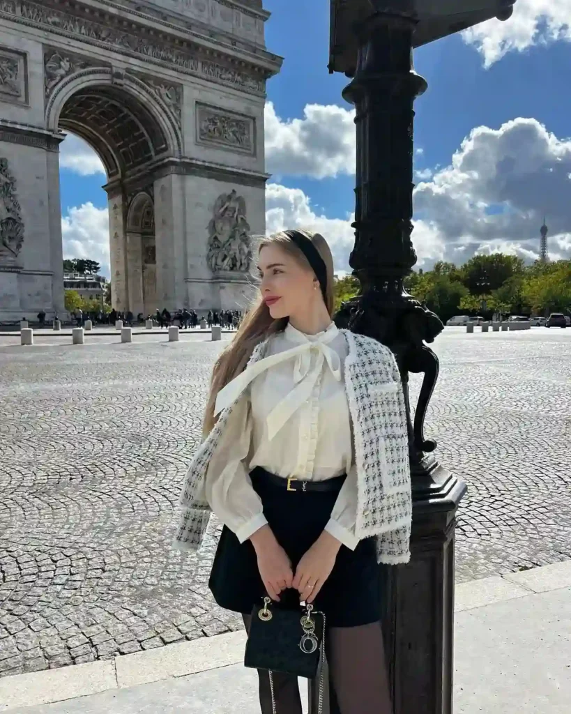Young woman in a stylish outfit poses at the Arc de Triomphe with blue skies and clouds in the background.