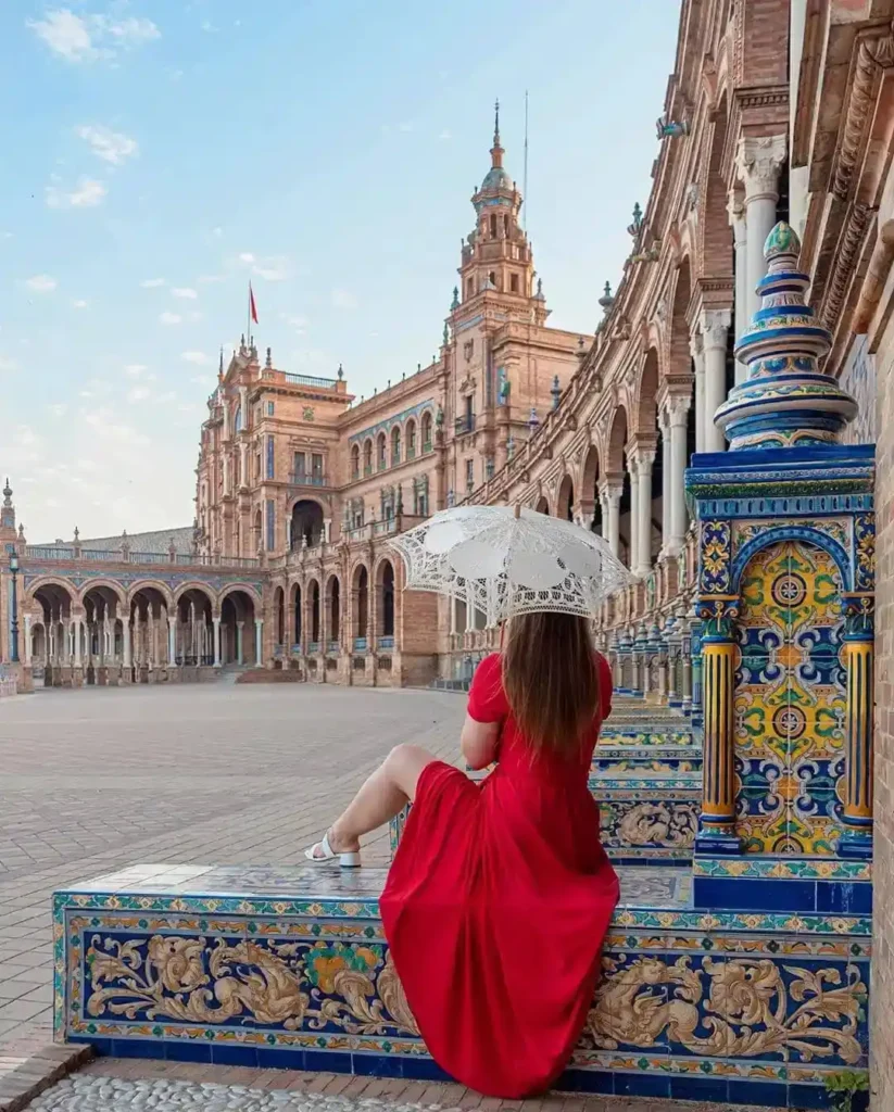 A woman in a red dress sits on a colorful tiled bench with a parasol at Plaza de España, Seville.