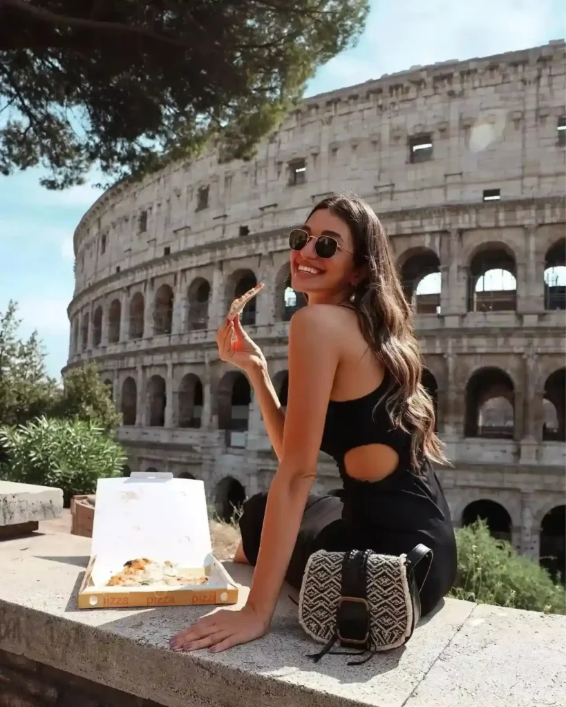 Woman enjoying pizza near the Colosseum in Rome, wearing sunglasses and a stylish black outfit.