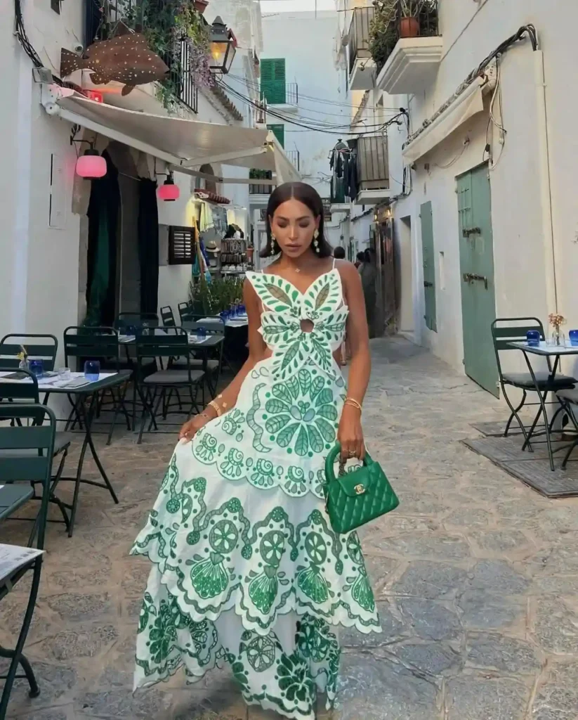 Woman in a green and white floral dress poses in a charming cobblestone street, surrounded by cafes and vibrant decor.