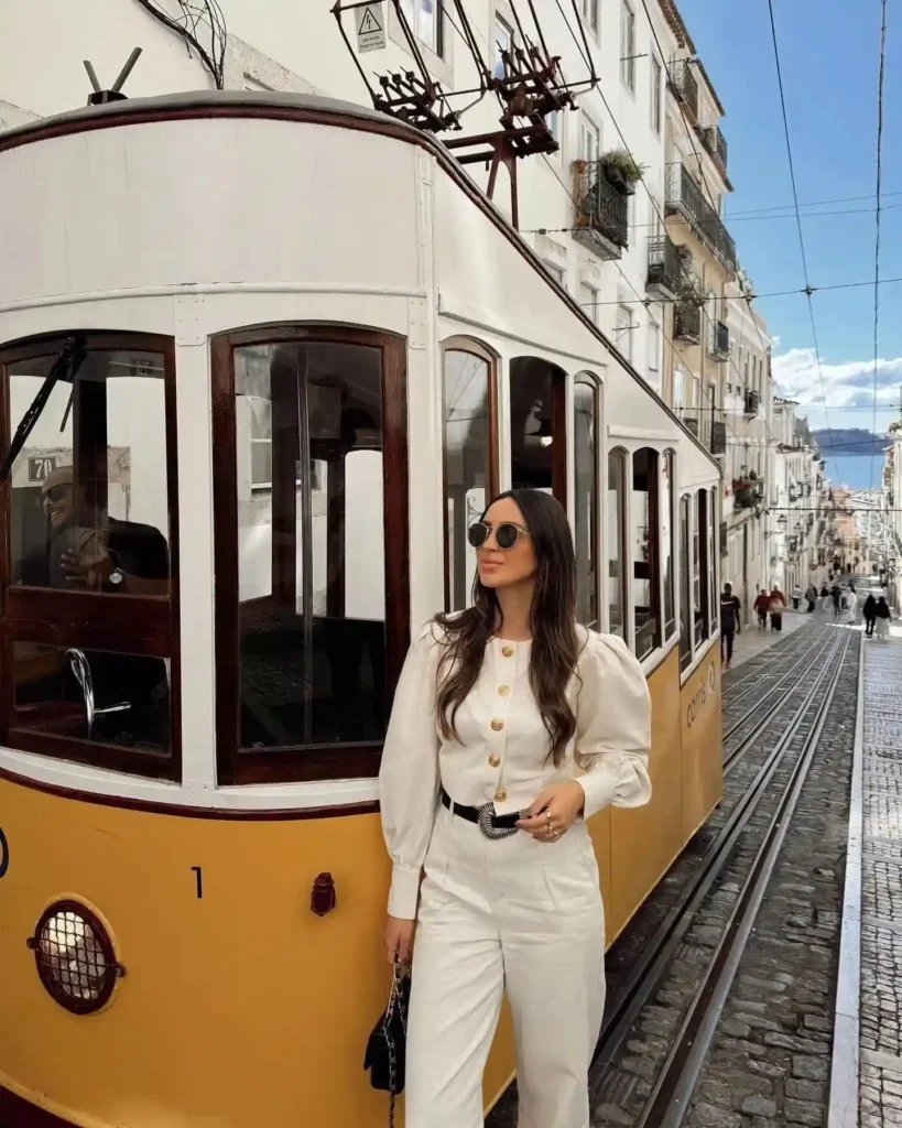 A woman in stylish white attire poses next to a vintage yellow tram in a picturesque street.