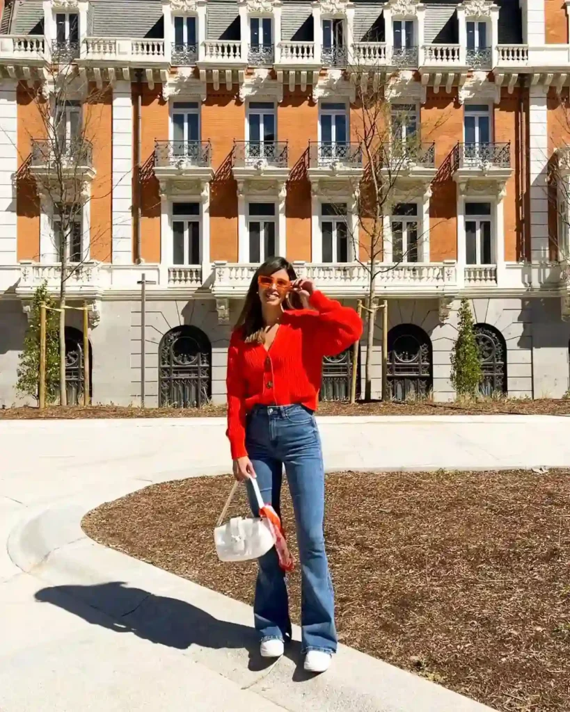 Woman in a red sweater and flared jeans poses in front of a stylish building, enjoying a sunny day outdoors.