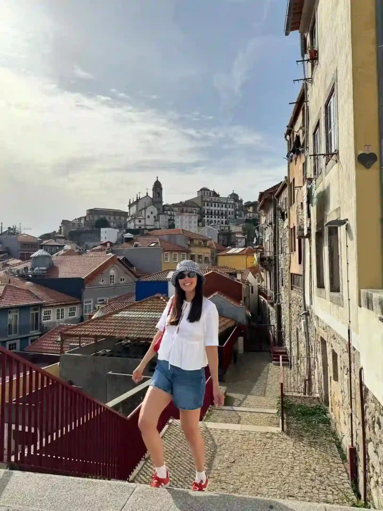 Young woman in casual summer attire smiles while walking down a scenic street with charming buildings in the background.